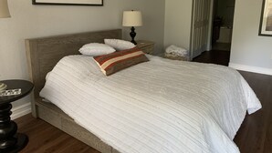 En pænt redt seng med hvidt sengetøj og en stribet pude, et natbord med en lampe, og et indrammet billede på væggen.