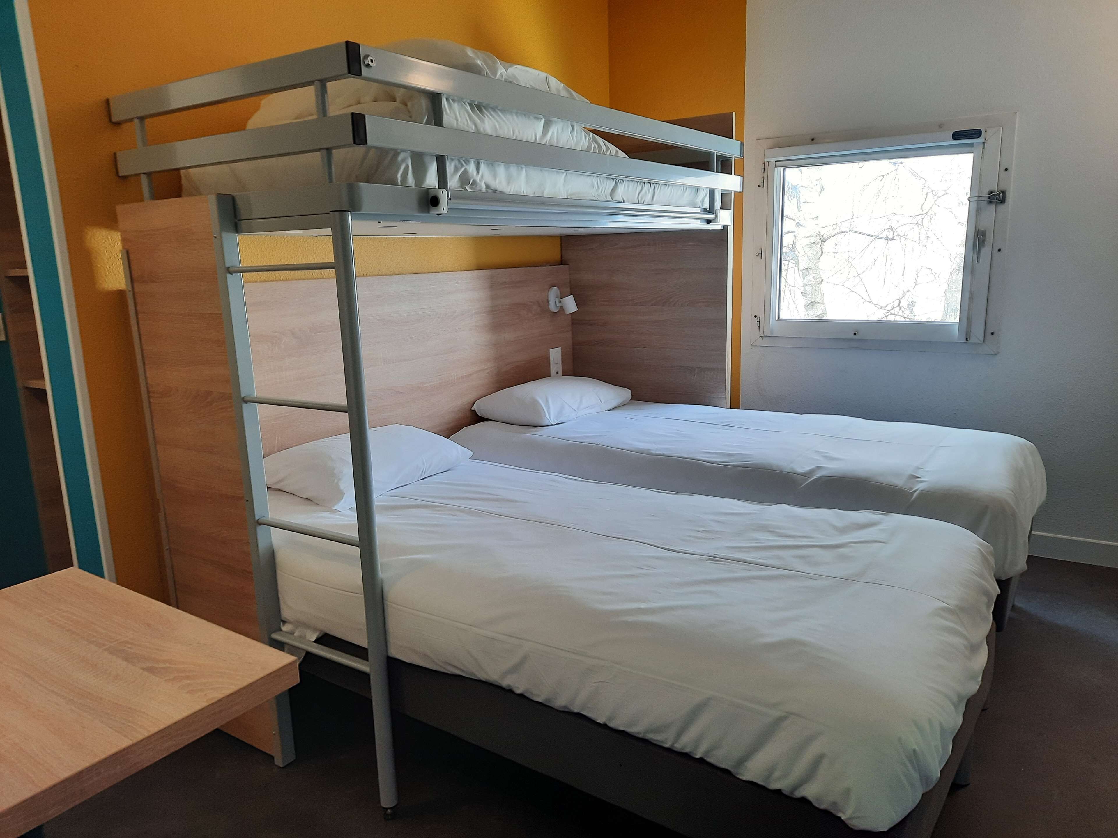 A bunk bed with two beds, a wooden desk, and a window with a view of trees.