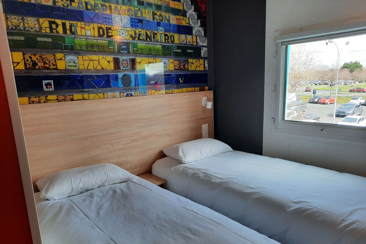 A room with two beds, a wooden headboard, a window with a view of a parking lot, and a colorful wall with various patterns and text.