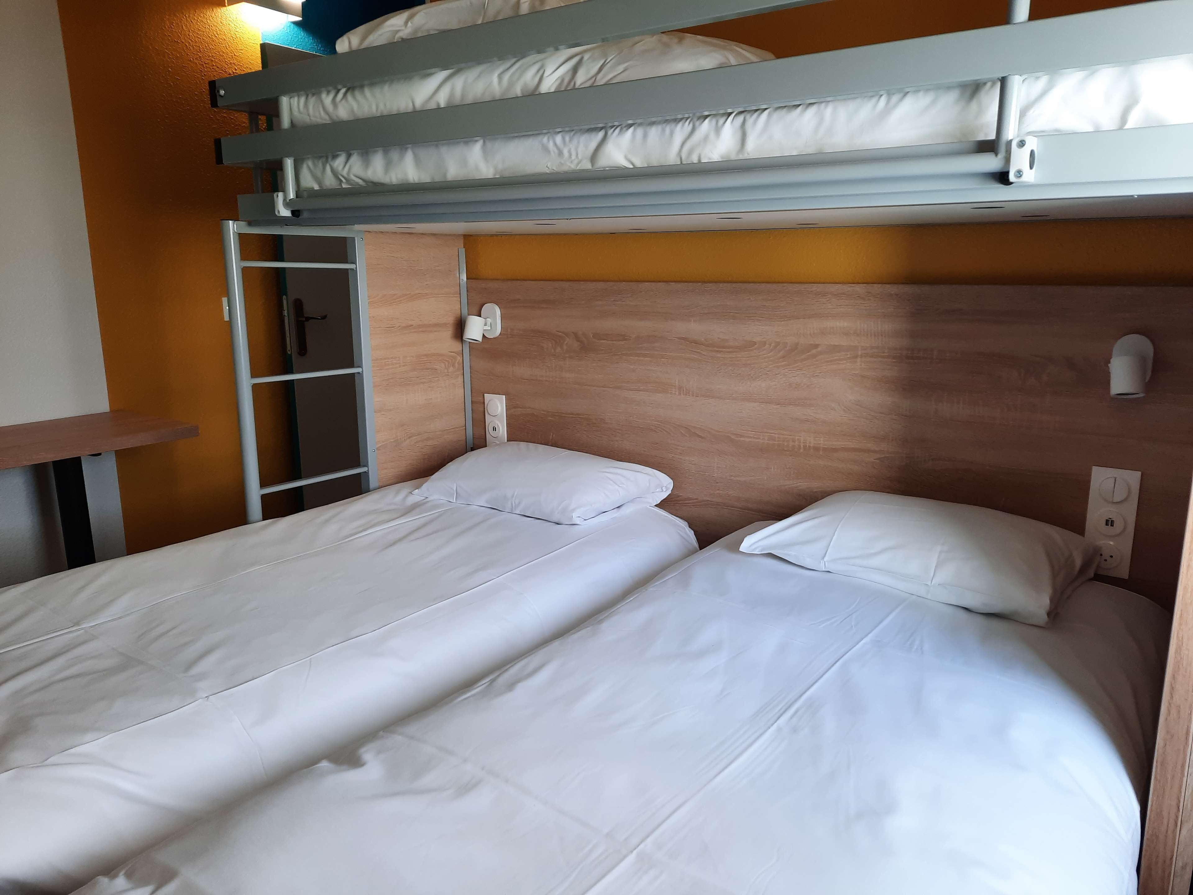 Un lit superposé avec deux lits simples en dessous, une tête de lit en bois, une lampe fixée au mur et une prise de courant.