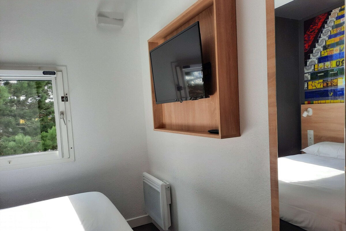A hotel room with two beds, a TV mounted on a wooden shelf, a window with a view of trees, and a radiator.
