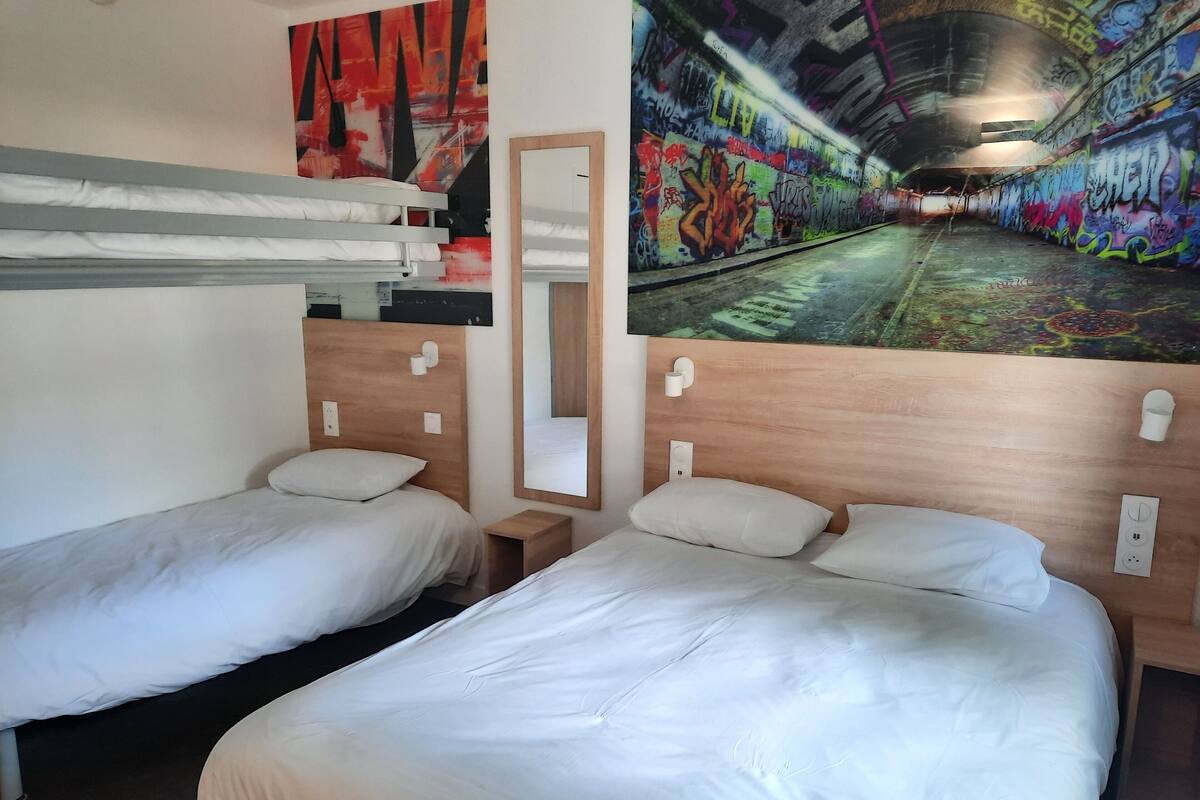 A hotel room with two single beds, a wooden headboard, a small bedside table, a mirror, and a wall mural of a graffiti-covered tunnel.
