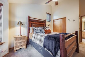 Quarto com cama de madeira, mesa de cabeceira com gavetas, guarda-roupa de madeira e ventoinha no teto.