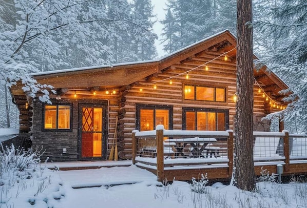 A log cabin with a wooden porch, a dining table, and a snow-covered landscape.