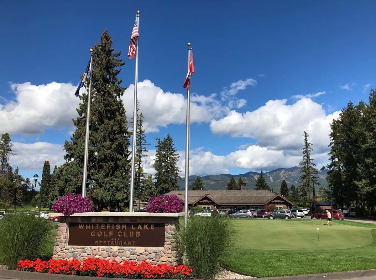 A golf club sign with flags, a mountain backdrop, and a building with a roof.