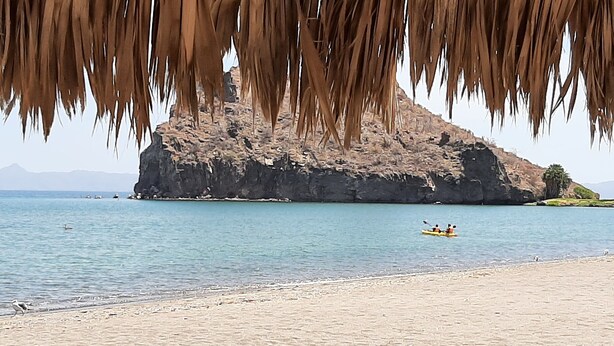A beach with a thatched roof structure, calm water, and a rocky hill in the background.