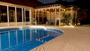 En swimmingpool med stenterasse, en overdækket patio med lyskæder, og en bygning i sten med store vinduer.
