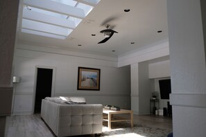 Un ampio soggiorno con soffitto alto, una ventola a soffitto, una grande finestra, un divano, un tavolino da caffè e un quadro appeso al muro.