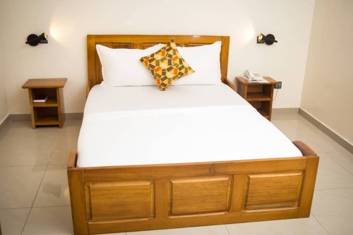 Una cama bien hecha con cabecera de madera, una mesita de noche con un teléfono y un pequeño estante con un libro.