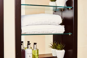Une salle de bain avec une étagère en verre où sont posés des serviettes pliées, une plante en pot et des articles de toilette.