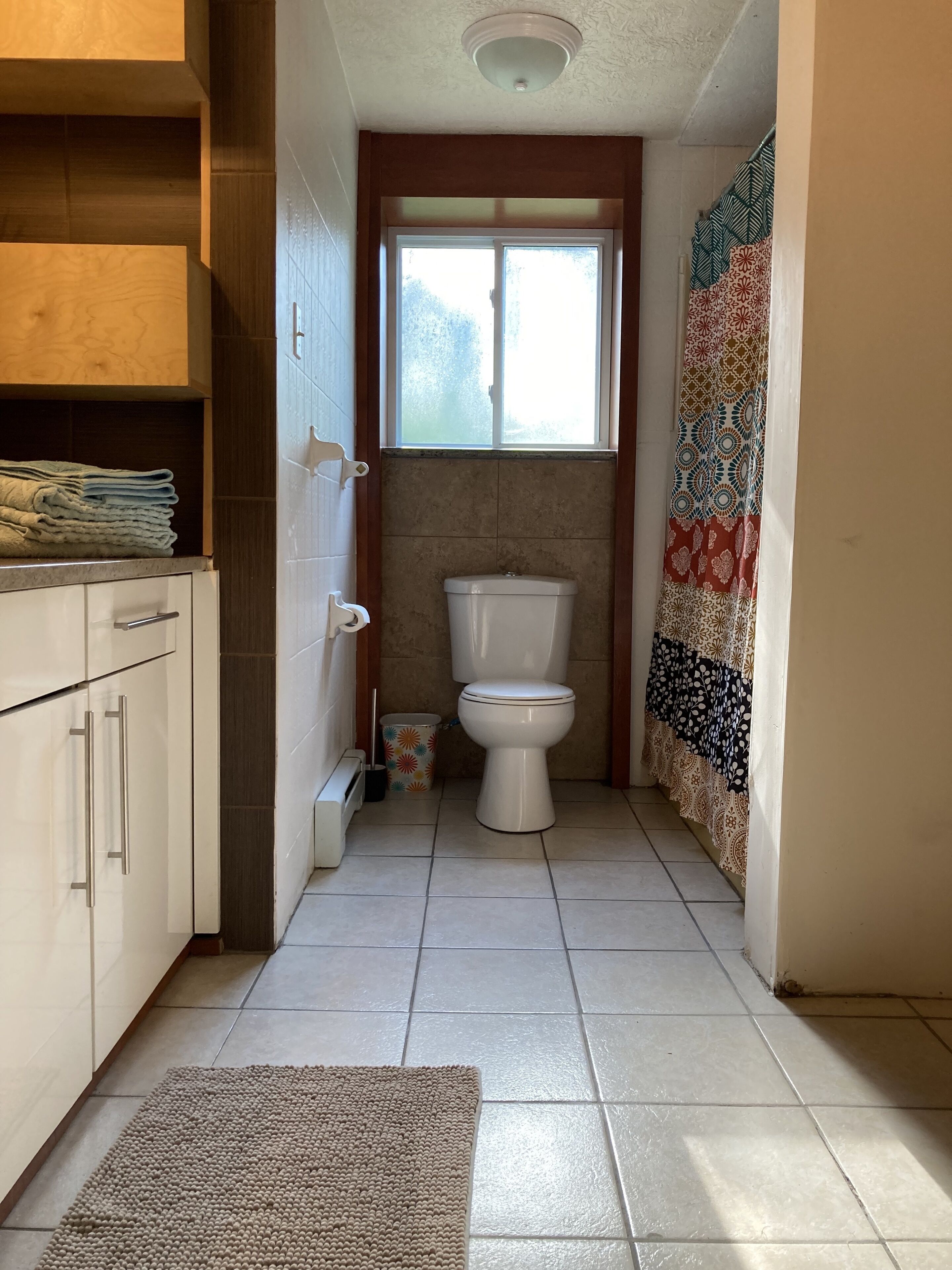 A narrow bathroom with a toilet, a window, and a patterned curtain.
