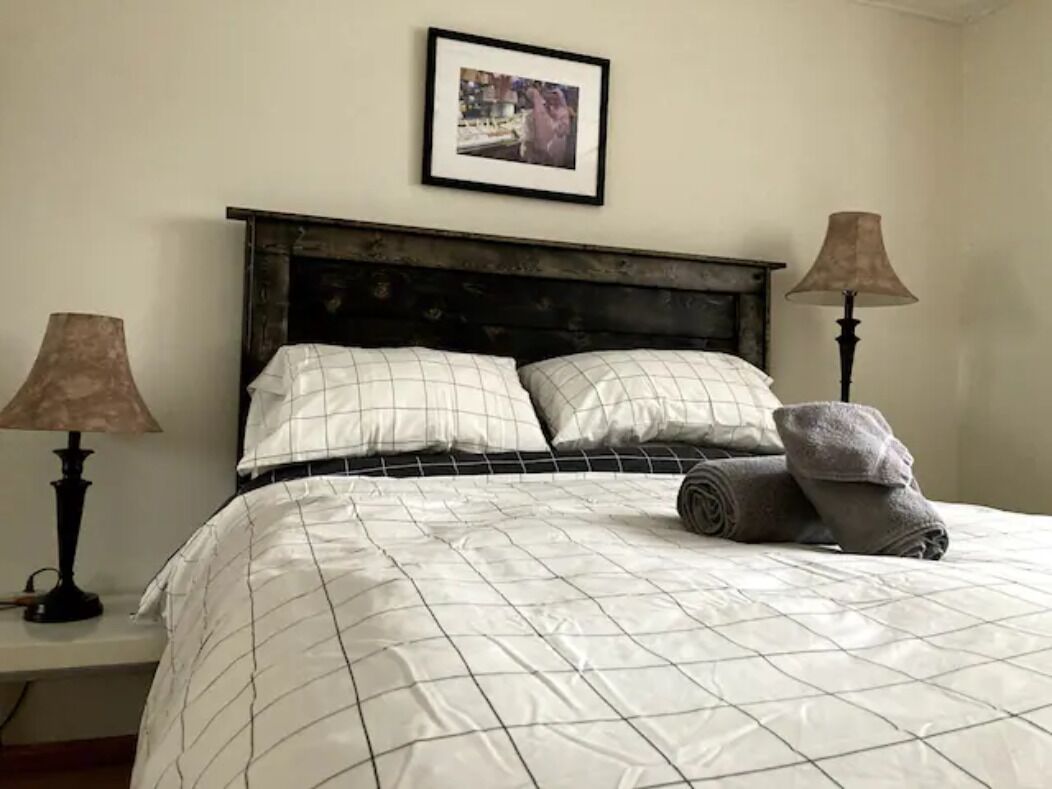 A neatly made bed with a checkered duvet, two bedside lamps, a framed picture on the wall, and a rolled towel on the bed.