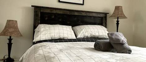 A neatly made bed with a checkered duvet, two bedside lamps, a framed picture on the wall, and a rolled towel on the bed.