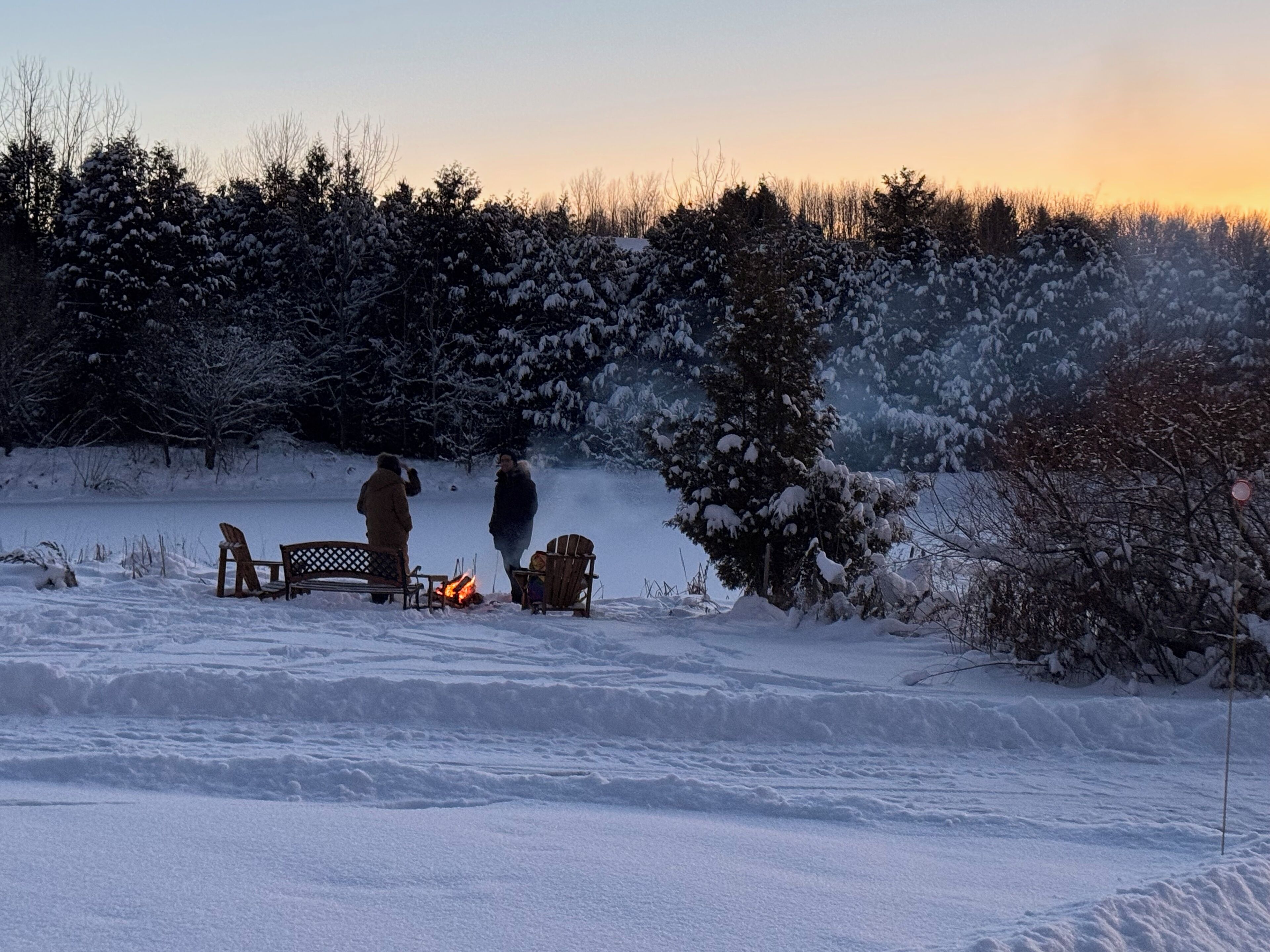 A snowy outdoor scene with a bench, a fire pit, and people gathered around.