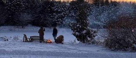 A snowy outdoor scene with a bench, a fire pit, and people gathered around.