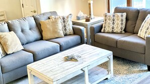 A living room with two grey sofas, a white coffee table, patterned cushions, a small side table with a lamp, and a view of the outdoors through a window.