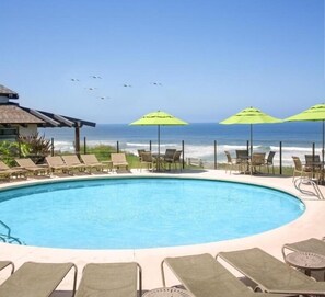 Área da piscina com espreguiçadeiras e guarda-sóis, com vista para o oceano.