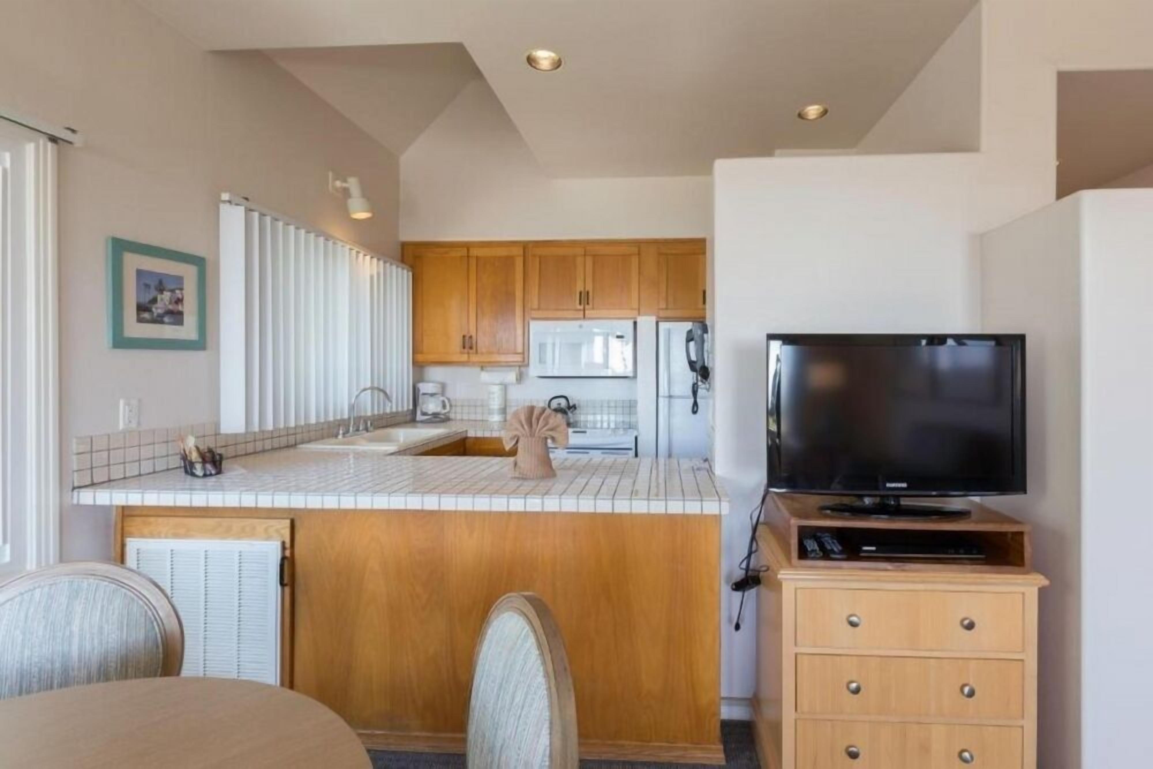 A compact kitchen with wooden cabinets, a microwave, a refrigerator, a sink, and a television on a wooden stand.