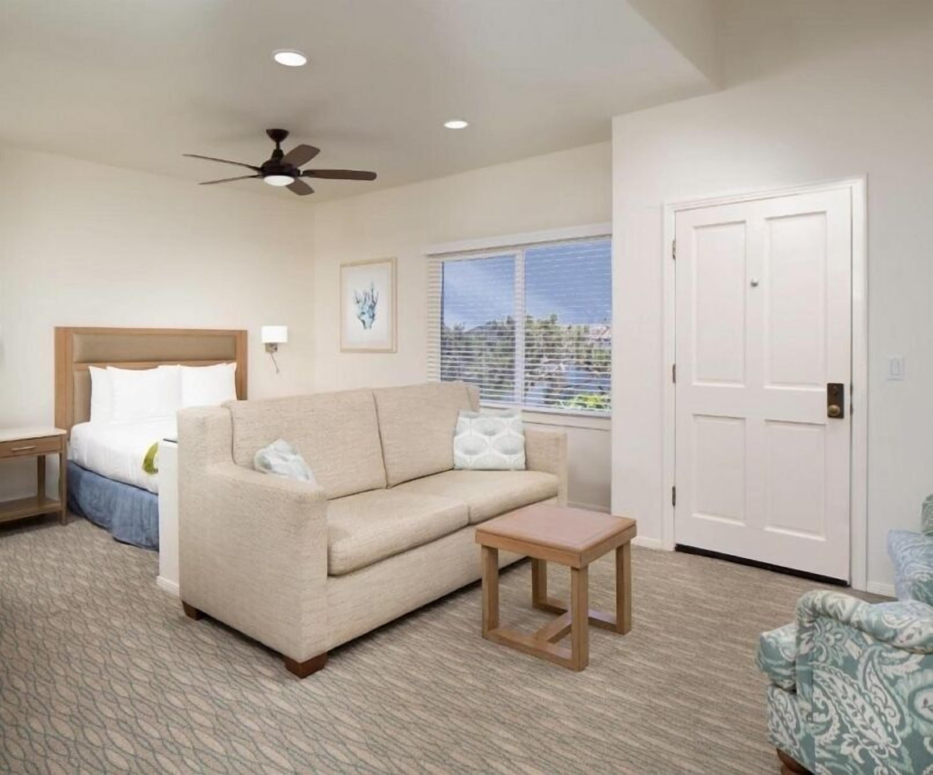 A hotel room with a bed, a sofa, a small table, a ceiling fan, and a window with blinds.