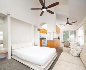 Quarto de hotel moderno com cama, sofá, cozinha compacta e dois ventiladores de teto.