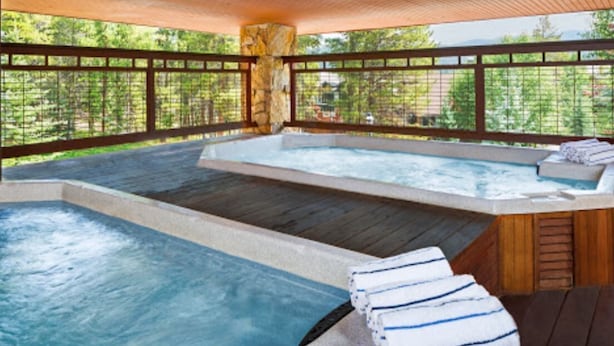 A hot tub area with a wooden deck, stone pillar, and a view of the surrounding forest.