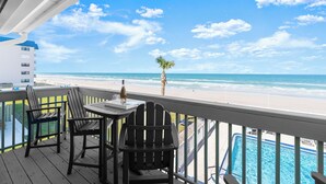 A balcony with a table and chairs overlooking a beach and a pool.