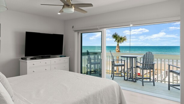 A bedroom with a TV, a bed, and a balcony with a view of the beach and ocean.