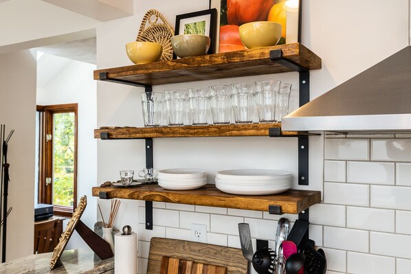 Cocina con estantes de madera que contienen cristalería y platos, una ventana con vista a follaje, y un salpicadero de azulejos.