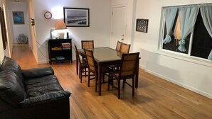 A dining area with a glass-top table and wooden chairs, a black leather sofa, a wall-mounted clock, a framed landscape painting, and a window with curtains.
