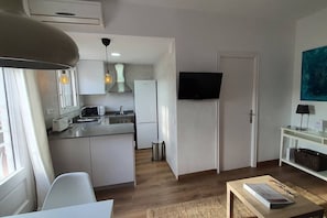 Et kompakt køkken med moderne hårde hvidevarer, et spiseområde med bord og stole, et fjernsyn monteret på væggen og et vindue med gardiner.