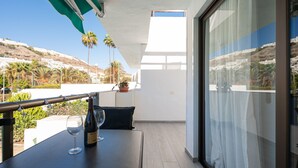 A balcony with a table, a wine glass, and a bottle of wine. There are palm trees and a view of a hillside in the background. - Sunny Angels by Canariasgetaway (Mogán)