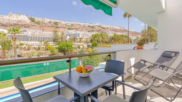 A balcony with a table, chairs, and a bowl of fruit. A pool is visible in the background. - Sunny Angels by Canariasgetaway (Mogán)