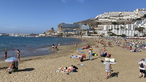 A beach with people sunbathing, swimming, and walking. There are umbrellas, beach chairs, and a building with multiple floors in the background. - Sunny Angels by Canariasgetaway (Mogán)