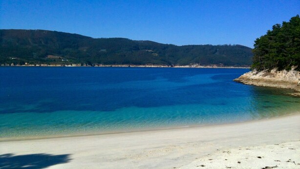 A beach with clear blue water, sandy shore, and forested hills in the background.