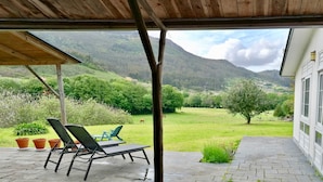 A covered patio with lounge chairs, a view of a green landscape, and a mountain in the background.