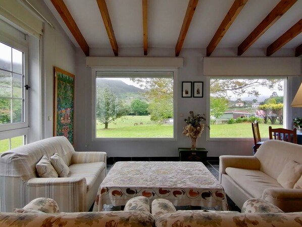 A cozy living room with two sofas, a coffee table, and large windows offering a view of a green landscape and mountains. - Beautiful, private villa with expansive garden close to beaches. (A Ponte de Mera)