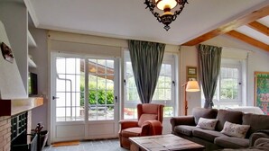 A living room with a fireplace, a sofa set, a coffee table, and a chair. There are two windows with curtains, and a chandelier hangs from the ceiling.