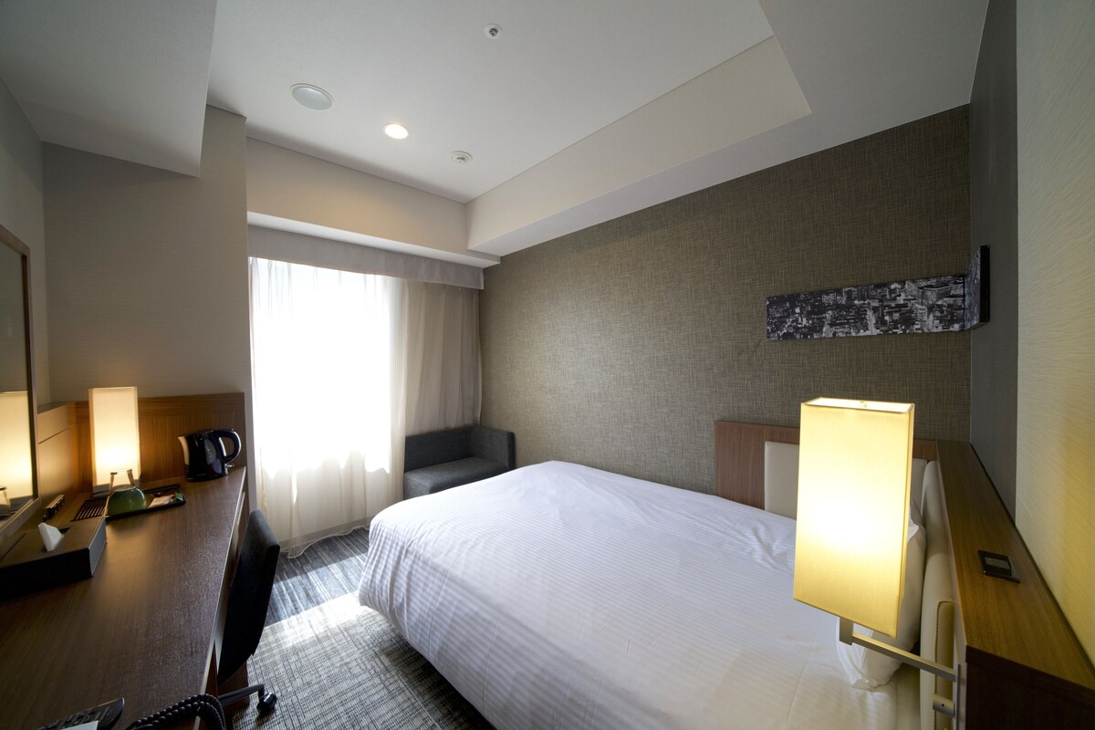 A hotel room with a bed, a desk with a lamp, a chair, and a large window with curtains.
