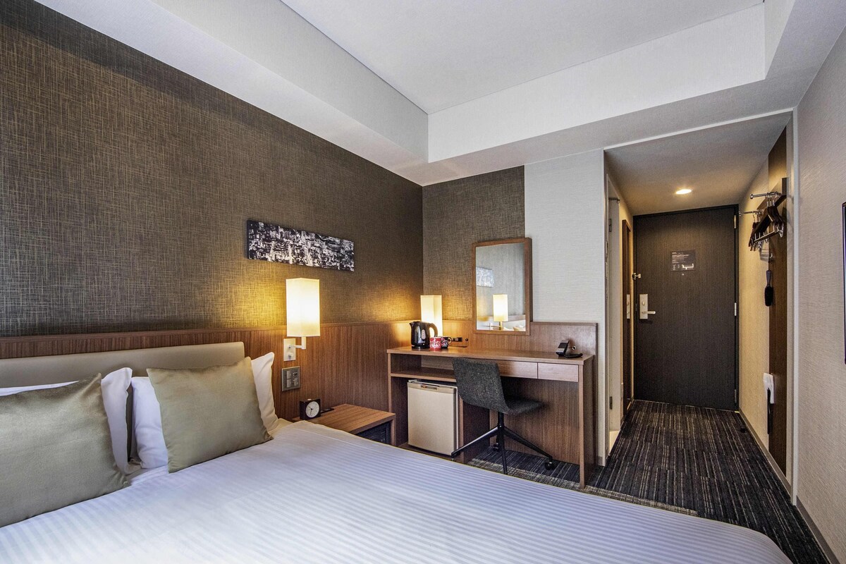 A hotel room with a large bed, a desk with a chair, a small refrigerator, a wall-mounted mirror, and a door leading to another room.