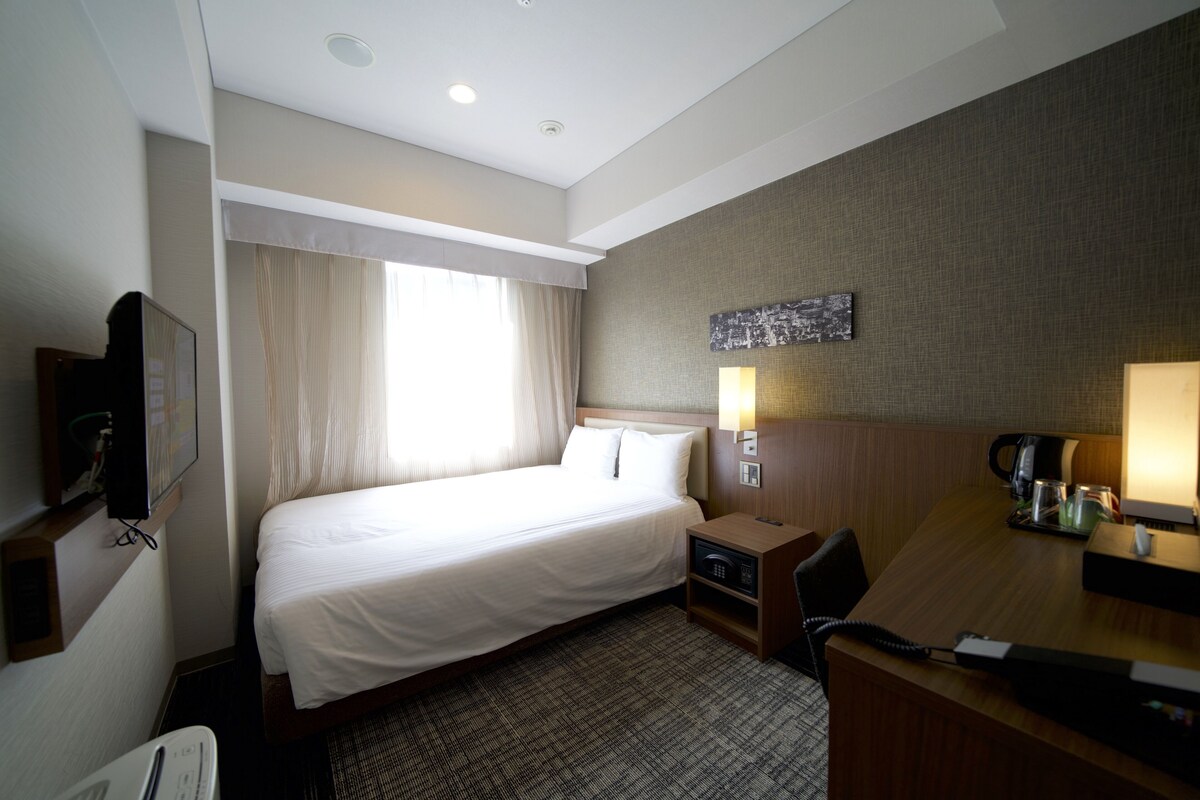 A hotel room with a bed, a desk, a chair, a TV, a lamp, and a coffee maker.