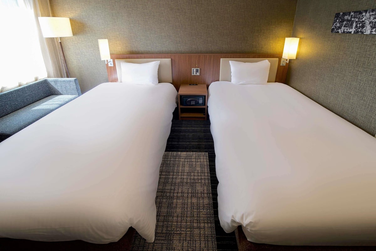 A hotel room with two beds, a nightstand, a lamp, and a view of the city.
