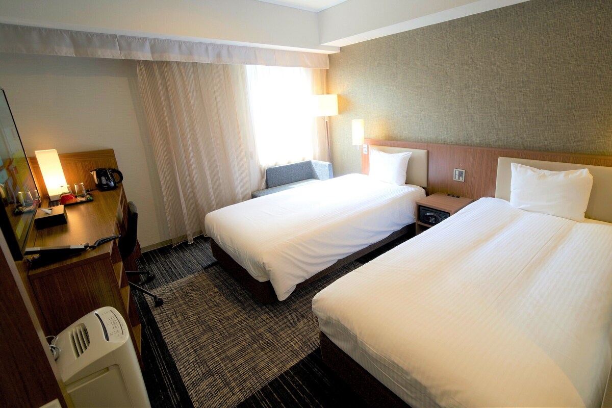 A hotel room with two beds, a desk, a chair, a lamp, and a window with curtains.