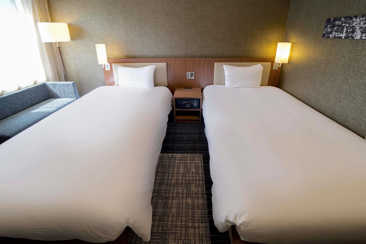 A hotel room with two beds, a nightstand, a lamp, and a view of the city.