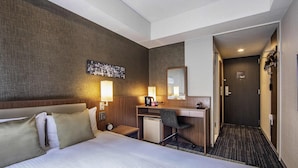 Habitación de hotel con una cama grande, un escritorio con silla, un refrigerador pequeño, un espejo montado en la pared y una puerta que da a otra habitación.