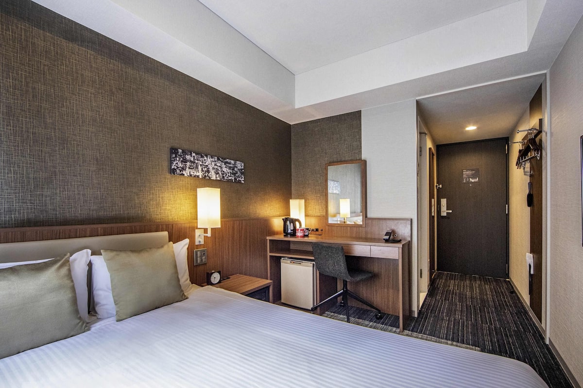 A hotel room with a large bed, a desk with a chair, a small refrigerator, a wall-mounted mirror, and a door leading to another room.