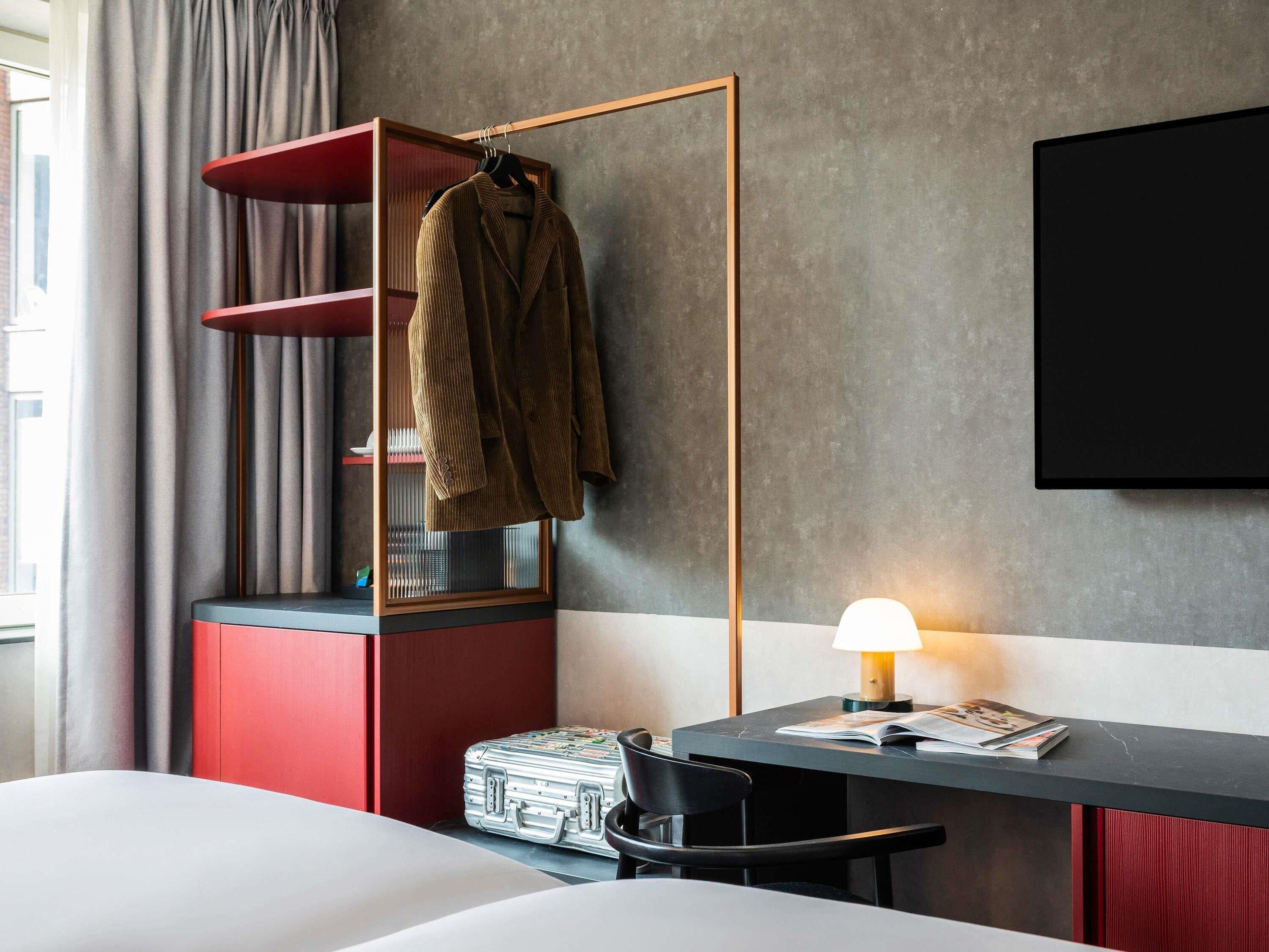 Une chambre d’hôtel avec un lit, un bureau doté d’une lampe, une chaise, une armoire rouge et une valise.