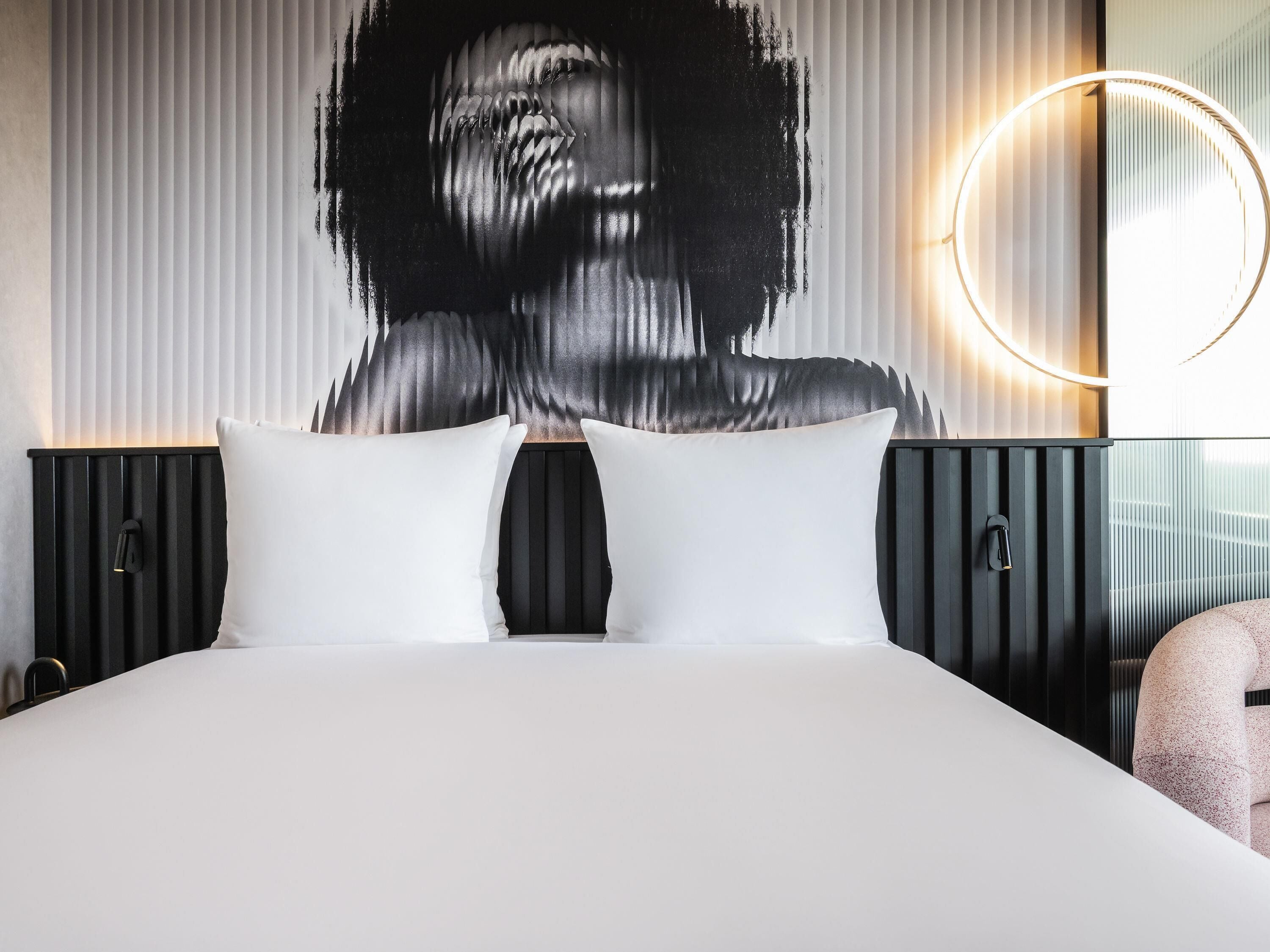 Habitación de hotel moderna con una cama grande, dos lámparas de noche, una luz circular montada en la pared y un cuadro abstracto de gran tamaño.
