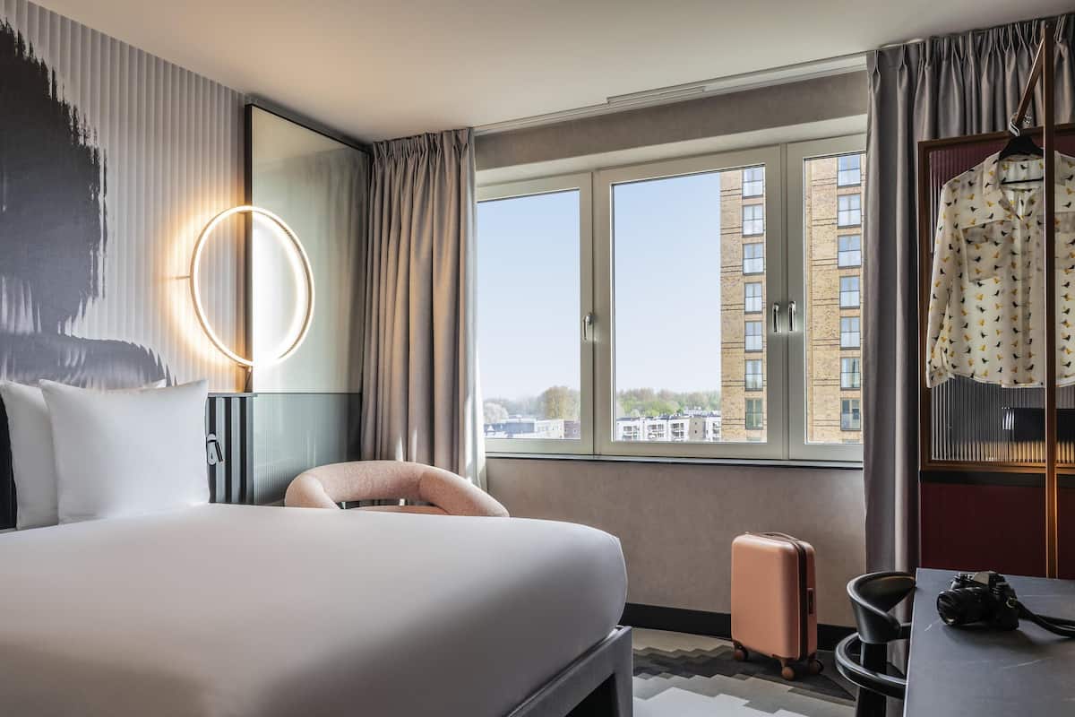 A hotel room with a bed, a chair, a suitcase, a table with a camera, a mirror, and a window with a view of buildings.