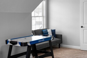 A room with a table hockey game, a grey sofa with blue cushions, a window with blinds, and a white door.