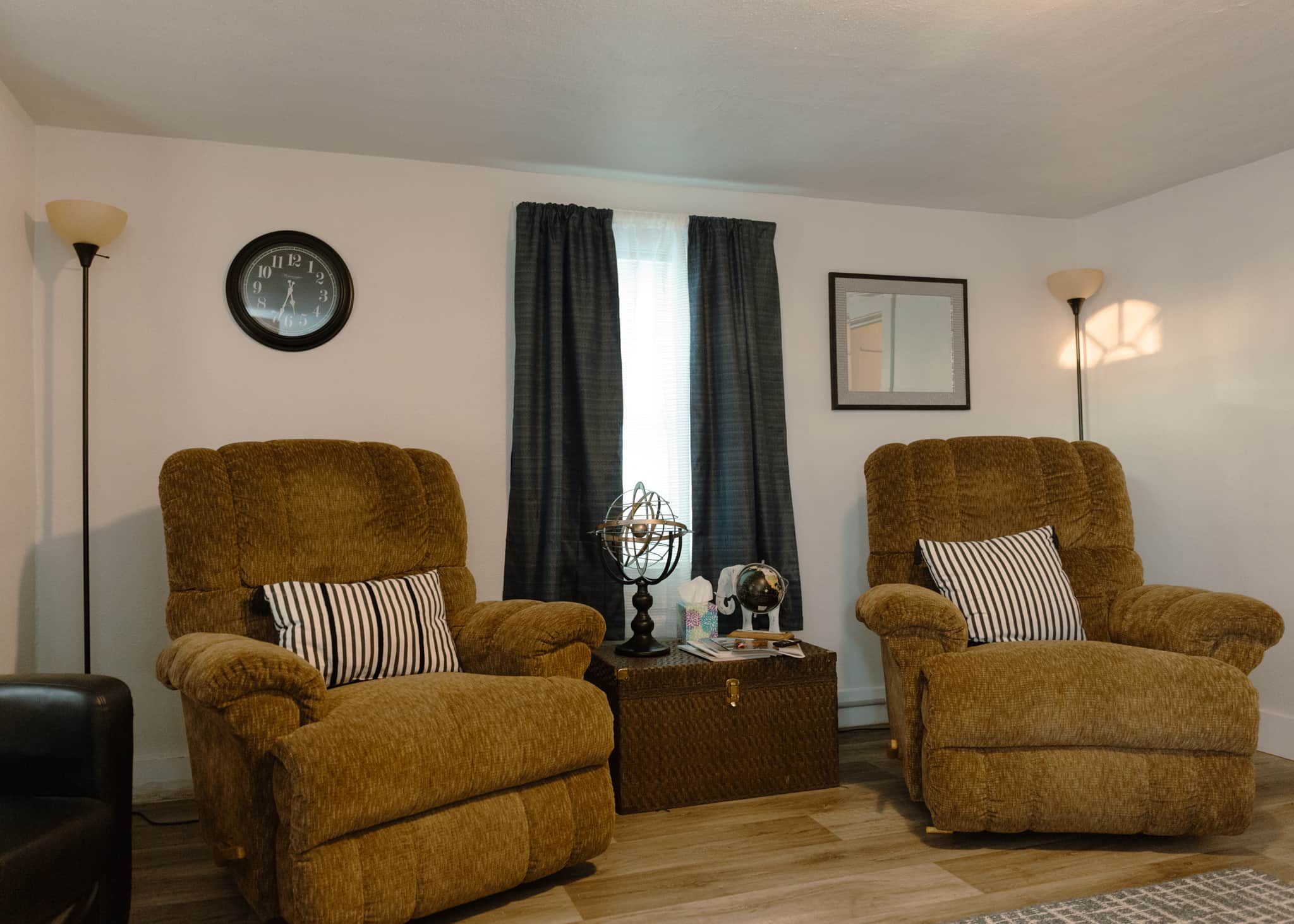 A living room with two brown armchairs, a small side table with a globe and books, a wall clock, and a framed picture.